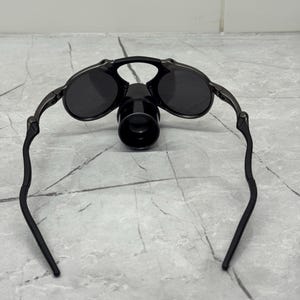 May include: Black rimmed sunglasses with dark lenses and a black, cylindrical attachment. The sunglasses have a sleek, modern design with curved temples. The lenses appear to be round and are set in a metal frame.
