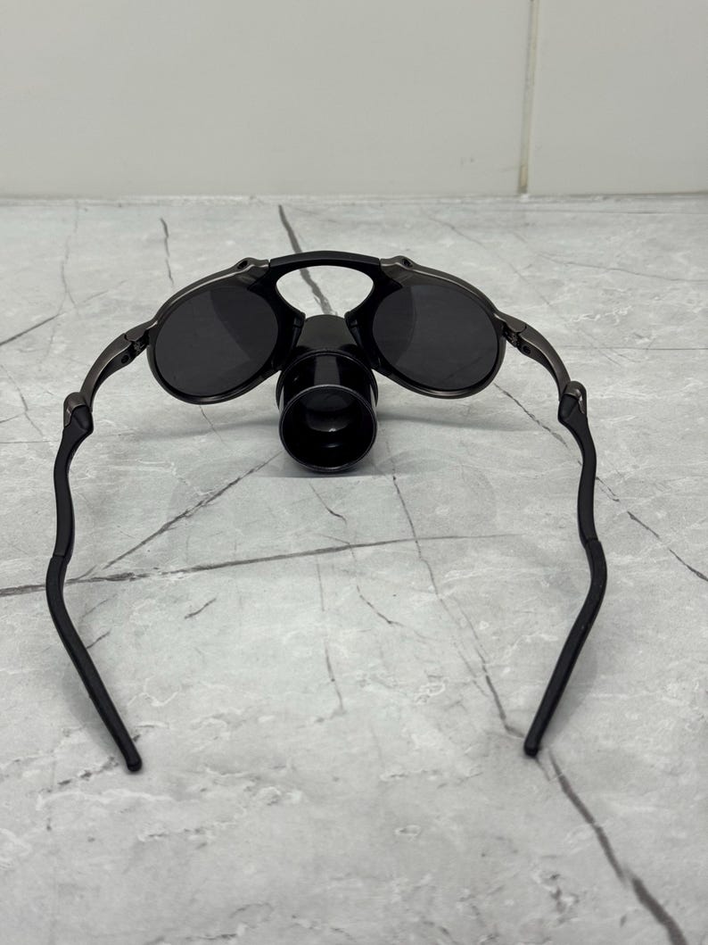 May include: Black rimmed sunglasses with dark lenses and a black, cylindrical attachment. The sunglasses have a sleek, modern design with curved temples. The lenses appear to be round and are set in a metal frame.