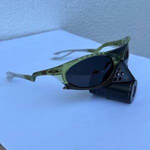 May include: Olive green and black sunglasses with a unique design. The sunglasses have a dark lens and a distinctive frame with a cylindrical attachment. The frame has a camouflage pattern.