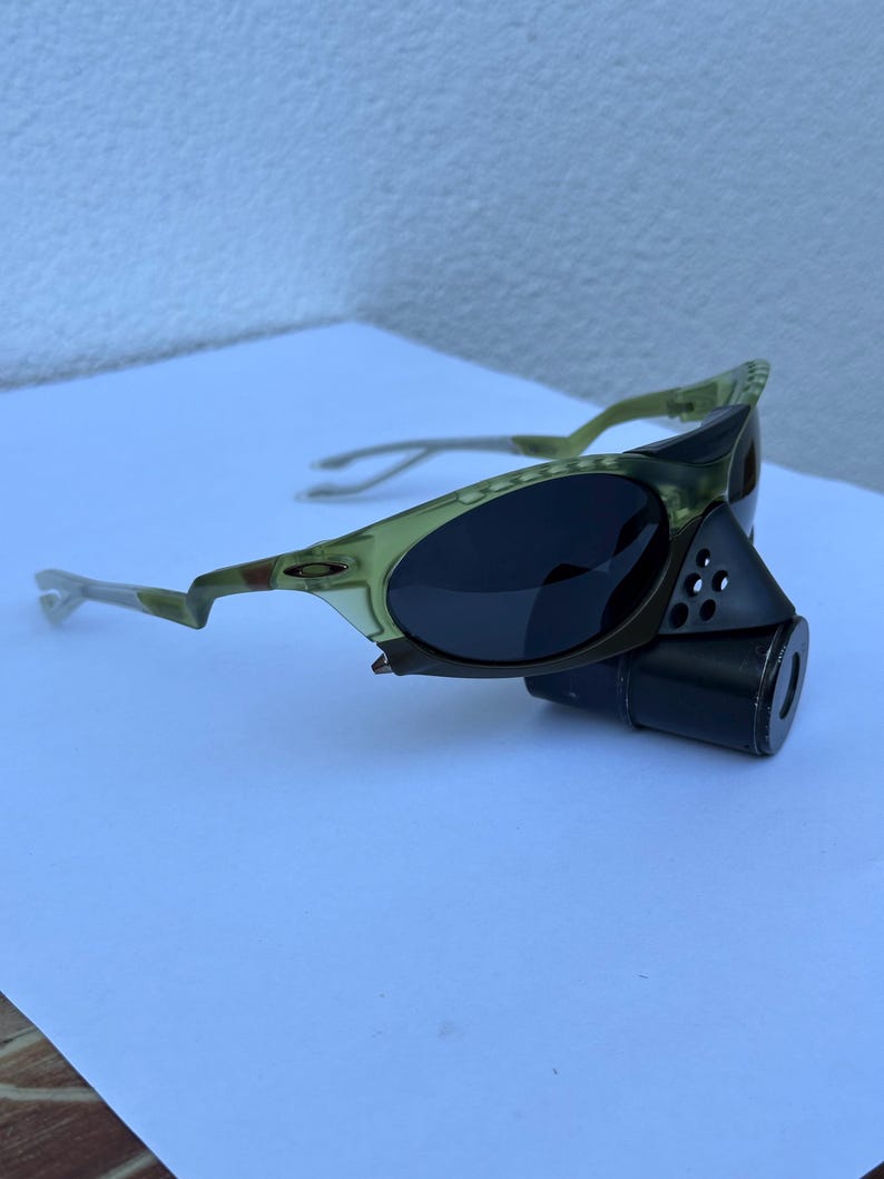 May include: Olive green and black sunglasses with a unique design. The sunglasses have a dark lens and a distinctive frame with a cylindrical attachment. The frame has a camouflage pattern.