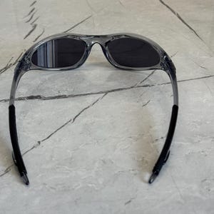 May include: Gray sunglasses with dark lenses. The frames are a translucent gray, and the temples are black. The sunglasses are designed with a curved shape for a sporty look.