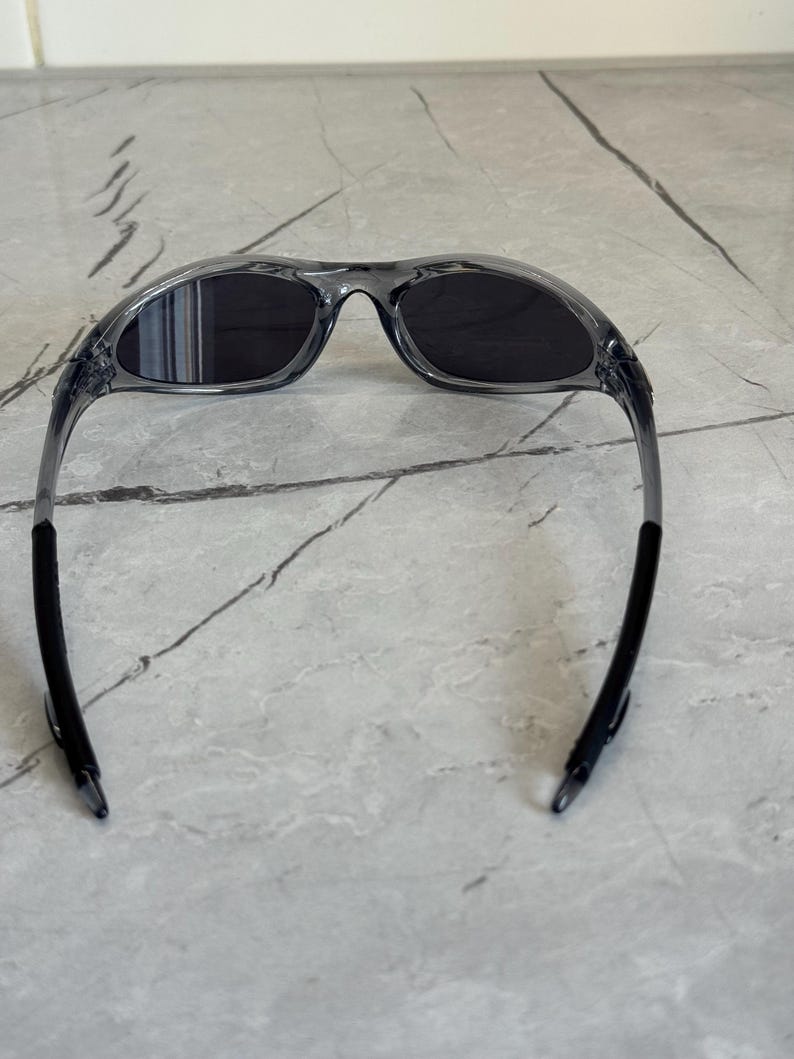 May include: Gray sunglasses with dark lenses. The frames are a translucent gray, and the temples are black. The sunglasses are designed with a curved shape for a sporty look.