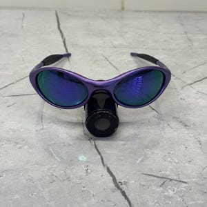 May include: Purple sunglasses with blue mirrored lenses. The sunglasses have a curved frame and black arms. The lenses reflect a blue and green hue. The sunglasses are resting on a black cylindrical object.