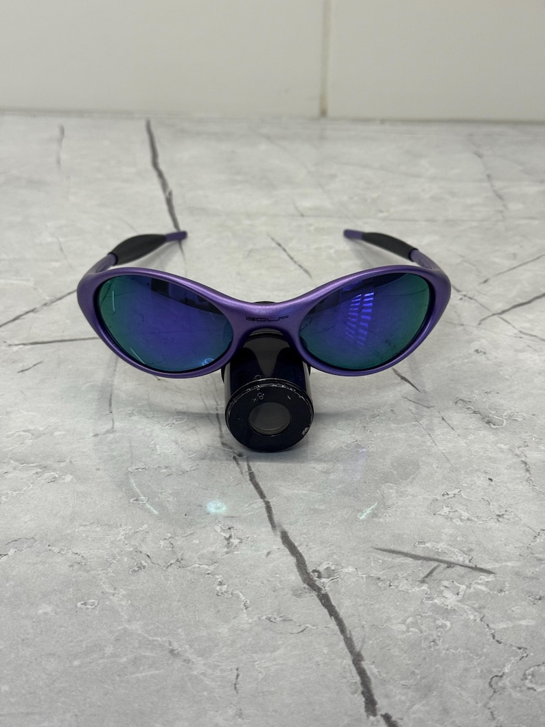 May include: Purple sunglasses with blue mirrored lenses. The sunglasses have a curved frame and black arms. The lenses reflect a blue and green hue. The sunglasses are resting on a black cylindrical object.