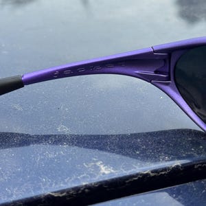 May include: Close-up of purple sunglasses with the word "JACKET" printed on the frame. The sunglasses have a sleek, modern design with dark lenses and a black arm.