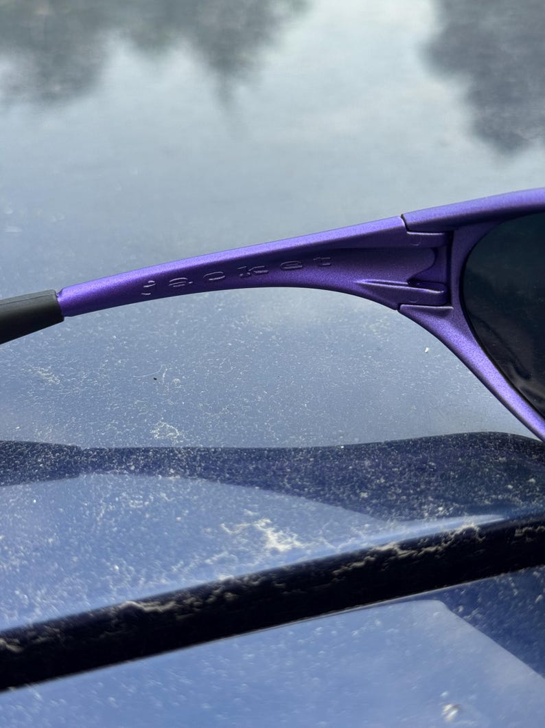 May include: Close-up of purple sunglasses with the word "JACKET" printed on the frame. The sunglasses have a sleek, modern design with dark lenses and a black arm.