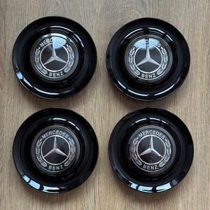 May include: Four black Mercedes-Benz wheel center caps. Each cap features the Mercedes-Benz logo, a silver three-pointed star within a laurel wreath, and the words "Mercedes Benz". The caps are glossy and circular.