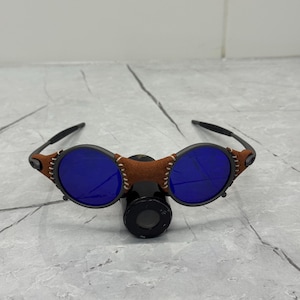 May include: Unique sunglasses with round blue lenses and a brown leather bridge. The frames are black with brown accents and white stitching. The sunglasses have a distinctive, retro design.