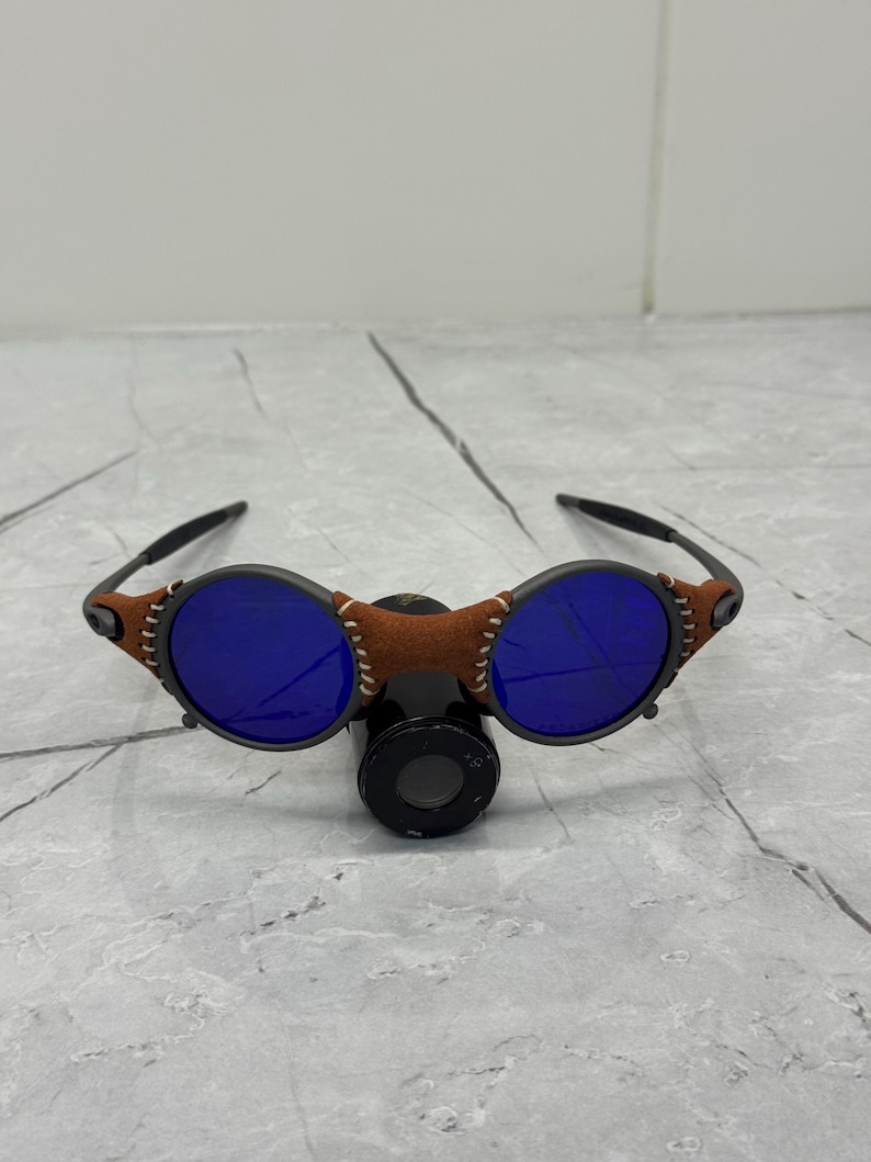 May include: Unique sunglasses with round blue lenses and a brown leather bridge. The frames are black with brown accents and white stitching. The sunglasses have a distinctive, retro design.