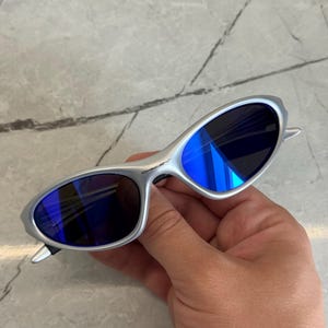 May include: Silver sunglasses with blue lenses. The oval lenses reflect light, creating a bright blue colour. The sunglasses have a minimalist design, with a thin frame and curved temples. A modern accessory for eye protection.