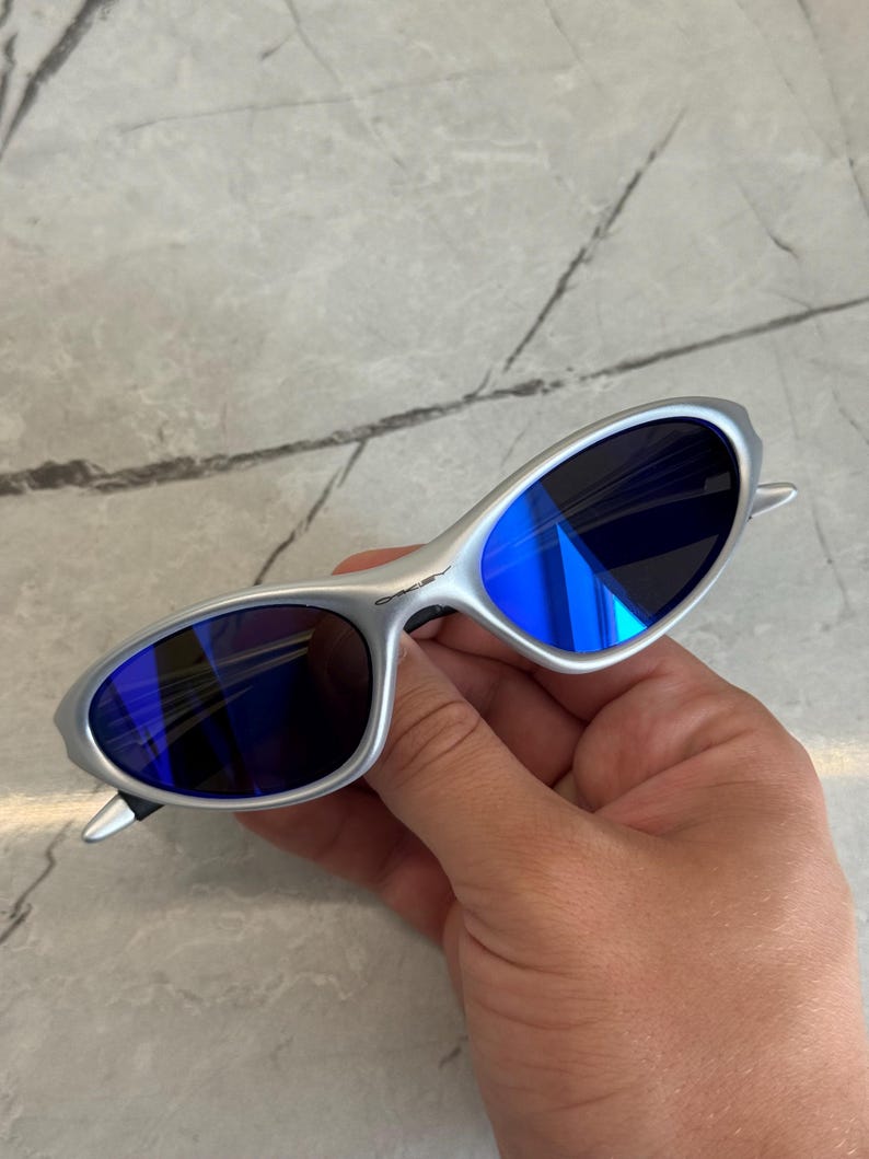 May include: Silver sunglasses with blue lenses. The oval lenses reflect light, creating a bright blue colour. The sunglasses have a minimalist design, with a thin frame and curved temples. A modern accessory for eye protection.