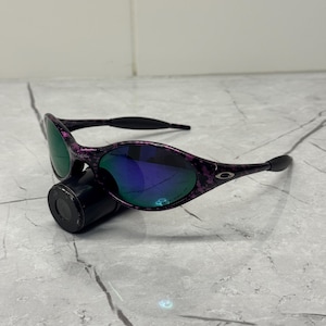 May include: Black and purple speckled sunglasses with mirrored lenses reflecting green and blue. The sunglasses are positioned on a black cylindrical object, set against a grey background.