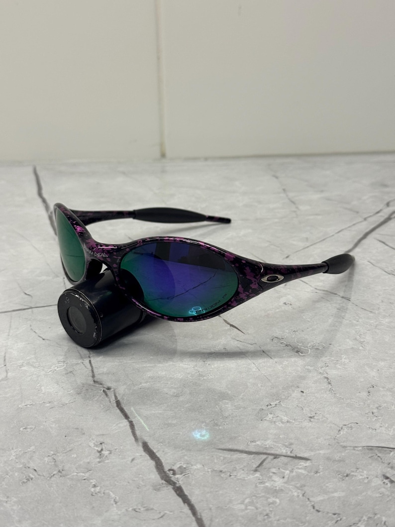 May include: Black and purple speckled sunglasses with mirrored lenses reflecting green and blue. The sunglasses are positioned on a black cylindrical object, set against a grey background.