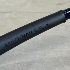 May include: Close-up of a black object with the embossed text "MADE IN U.S.A." The object has a smooth, matte finish and appears to be a handle or part of a larger tool. The background is a light gray, textured surface.