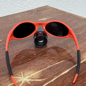 Pu&ograve; includere: Occhiali da sole rossi con lenti nere e terminali in gomma nera sulle aste. Gli occhiali da sole sono appoggiati su una superficie di legno. Design sportivo e avvolgente.