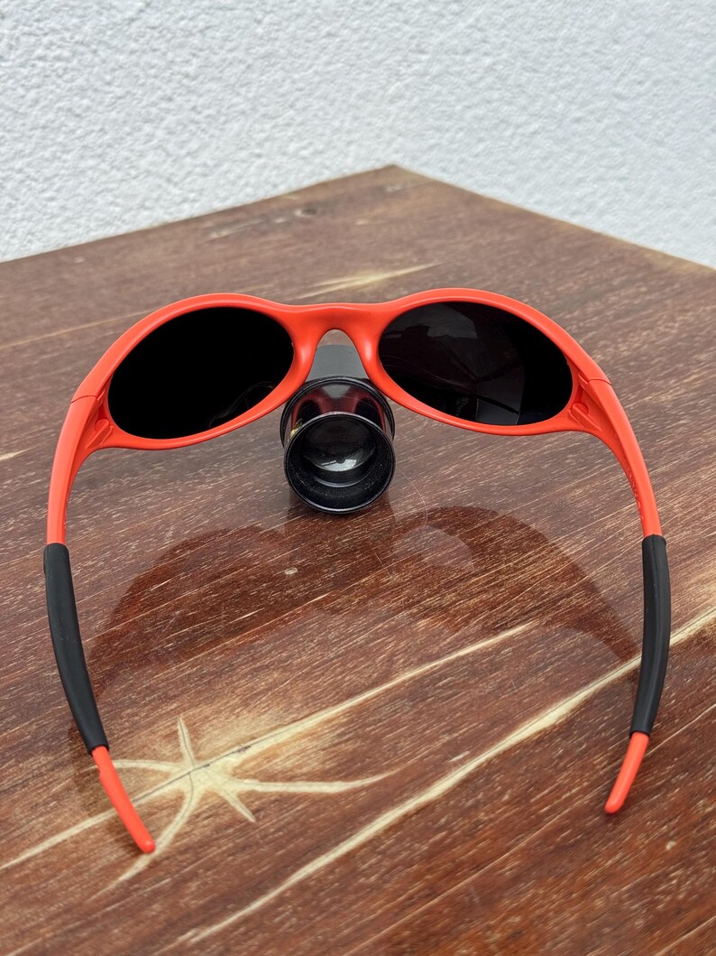 Pu&ograve; includere: Occhiali da sole rossi con lenti nere e terminali in gomma nera sulle aste. Gli occhiali da sole sono appoggiati su una superficie di legno. Design sportivo e avvolgente.