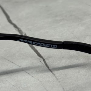 May include: Close-up of a black eyeglass arm with the word "MADMAN" printed in gray. The arm is curved and appears to be made of a matte material. The background is a light gray, textured surface.