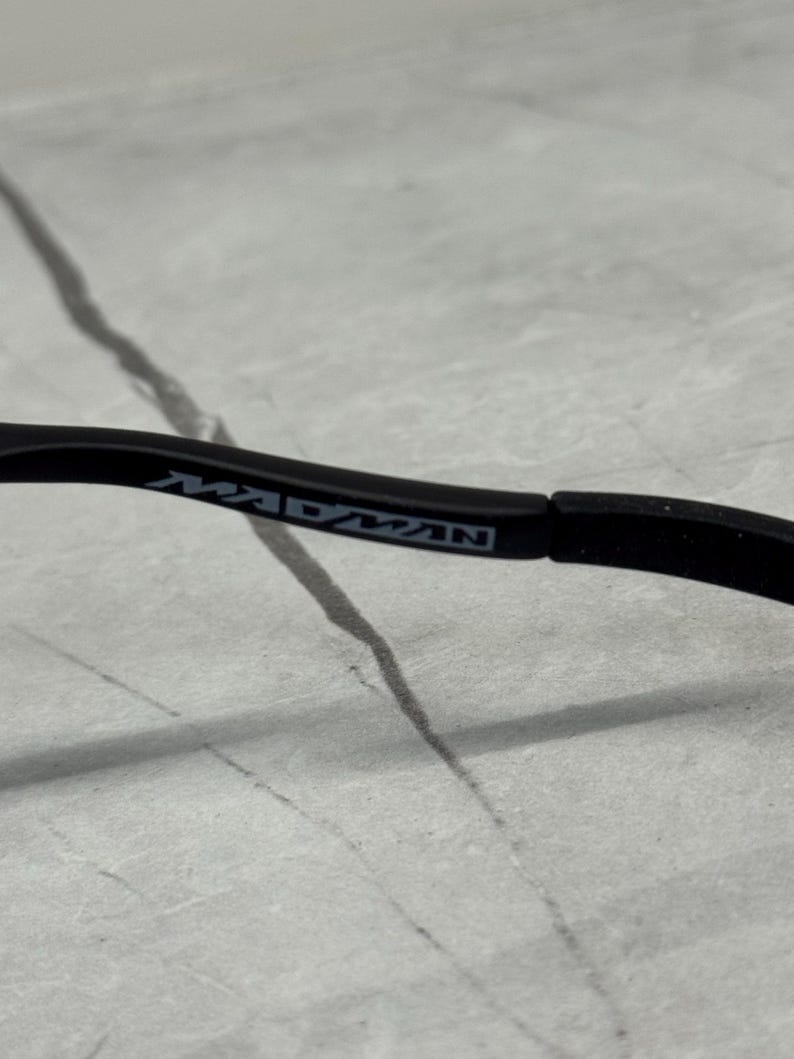 May include: Close-up of a black eyeglass arm with the word "MADMAN" printed in gray. The arm is curved and appears to be made of a matte material. The background is a light gray, textured surface.