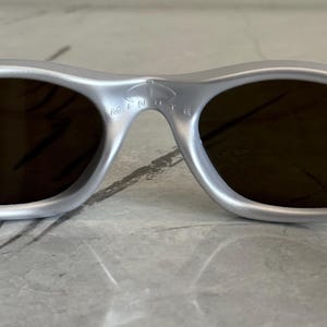 May include: Silver sunglasses with dark lenses. The sunglasses have a curved frame and the word "MINUTE" is printed on the bridge. The lenses are a dark brown colour.