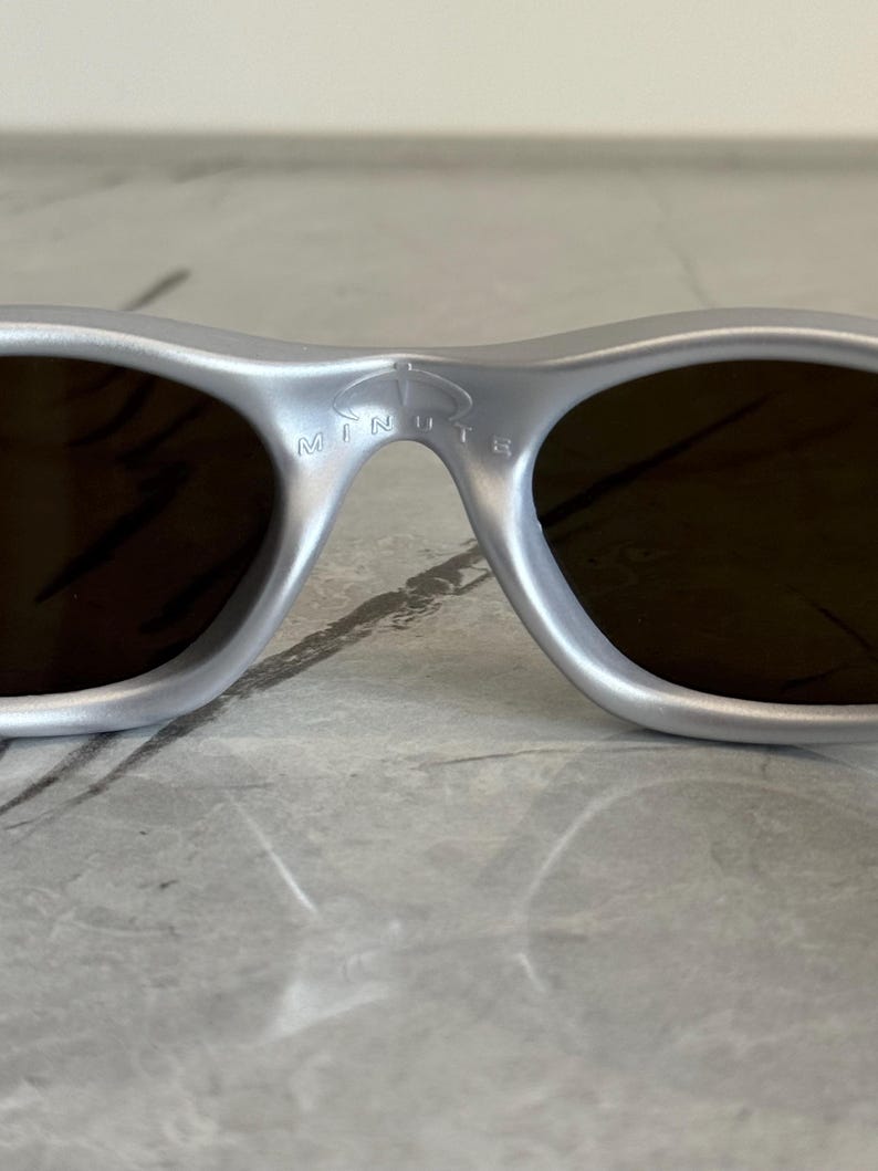 May include: Silver sunglasses with dark lenses. The sunglasses have a curved frame and the word "MINUTE" is printed on the bridge. The lenses are a dark brown colour.