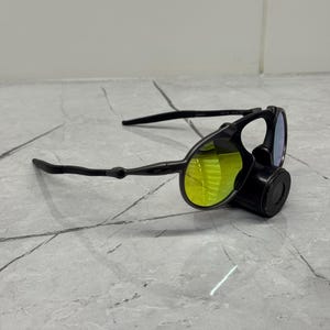 May include: Black sunglasses with yellow-tinted lenses and a unique design. The sunglasses have a distinctive shape with a nose piece and sleek black arms. The lenses appear to be reflective, and the overall style is modern and sporty.