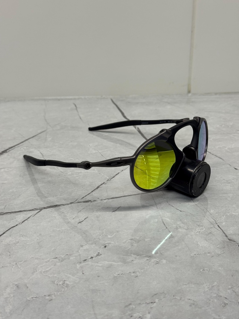May include: Black sunglasses with yellow-tinted lenses and a unique design. The sunglasses have a distinctive shape with a nose piece and sleek black arms. The lenses appear to be reflective, and the overall style is modern and sporty.