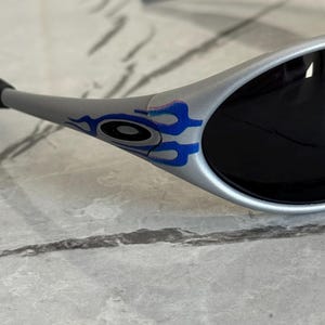 Puede incluir: Gafas de sol plateadas con lentes negras. Las patillas presentan un dise&ntilde;o de llamas azules y moradas. Las gafas de sol est&aacute;n sobre un fondo gris claro.