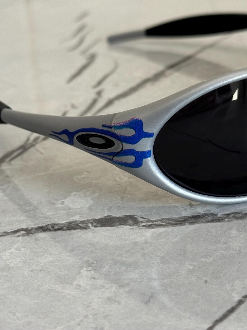 Puede incluir: Gafas de sol plateadas con lentes negras. Las patillas presentan un dise&ntilde;o de llamas azules y moradas. Las gafas de sol est&aacute;n sobre un fondo gris claro.