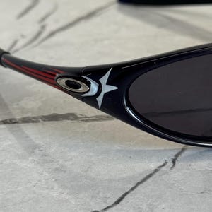 K&ouml;nnte beinhalten: Schwarze Sonnenbrille mit Sternmuster am B&uuml;gel. Die Sonnenbrille hat einen schwarzen Rahmen mit roten und wei&szlig;en Akzenten. Die Gl&auml;ser sind dunkel, und das Design wirkt sportlich und modern.