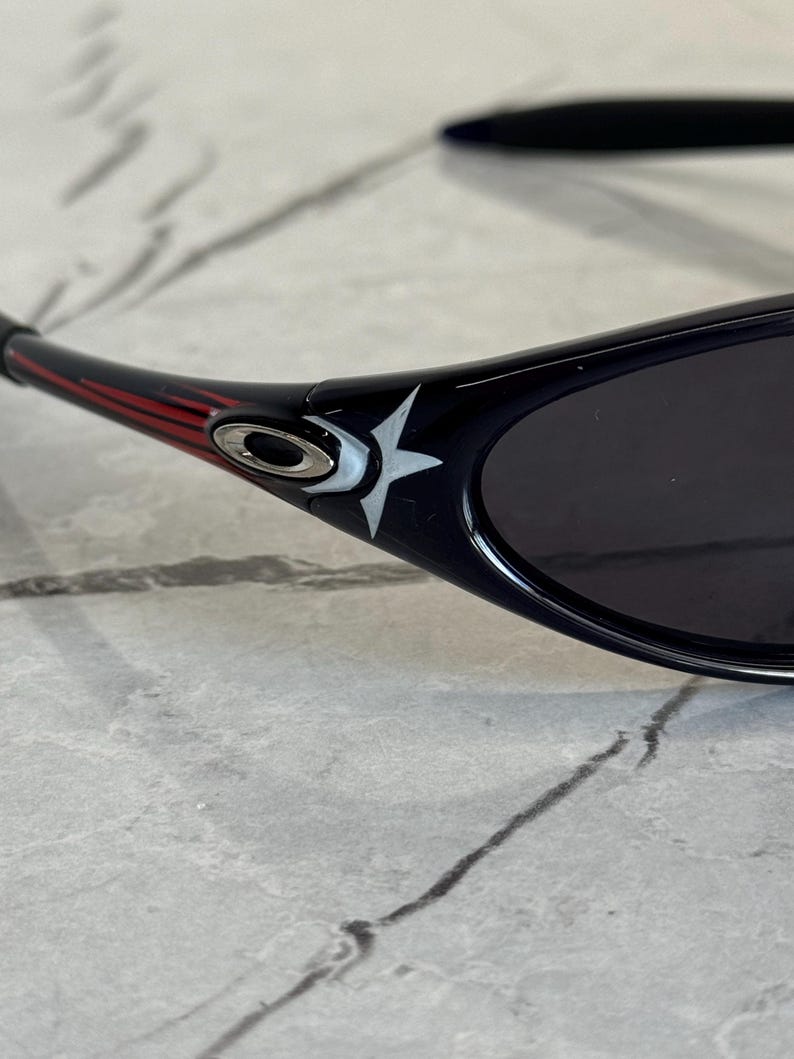 K&ouml;nnte beinhalten: Schwarze Sonnenbrille mit Sternmuster am B&uuml;gel. Die Sonnenbrille hat einen schwarzen Rahmen mit roten und wei&szlig;en Akzenten. Die Gl&auml;ser sind dunkel, und das Design wirkt sportlich und modern.