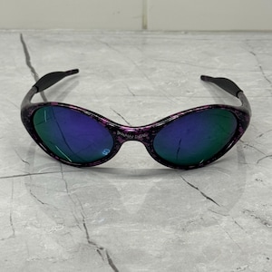May include: Oval sunglasses with a black and purple speckled frame and blue-green lenses. The sunglasses have a curved design. The text "BRAIN DEAD" is visible on the frame. The background is marble.
