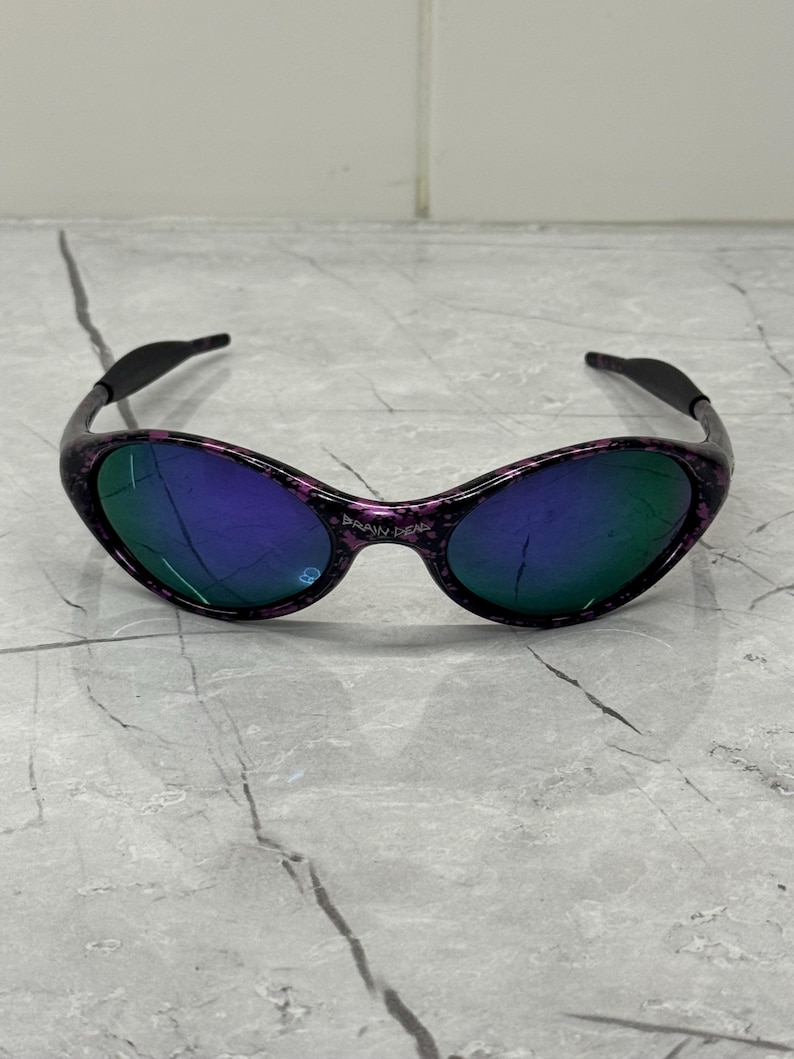 May include: Oval sunglasses with a black and purple speckled frame and blue-green lenses. The sunglasses have a curved design. The text "BRAIN DEAD" is visible on the frame. The background is marble.