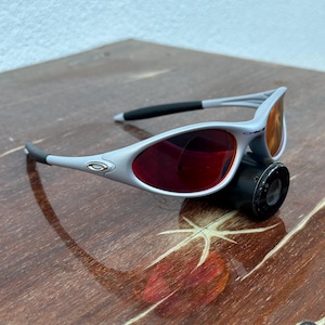 May include: Gray and black sunglasses with red lenses. The sunglasses have a sleek, modern design with a silver logo on the temples. The lenses are a vibrant red color, and the frames are a light gray.