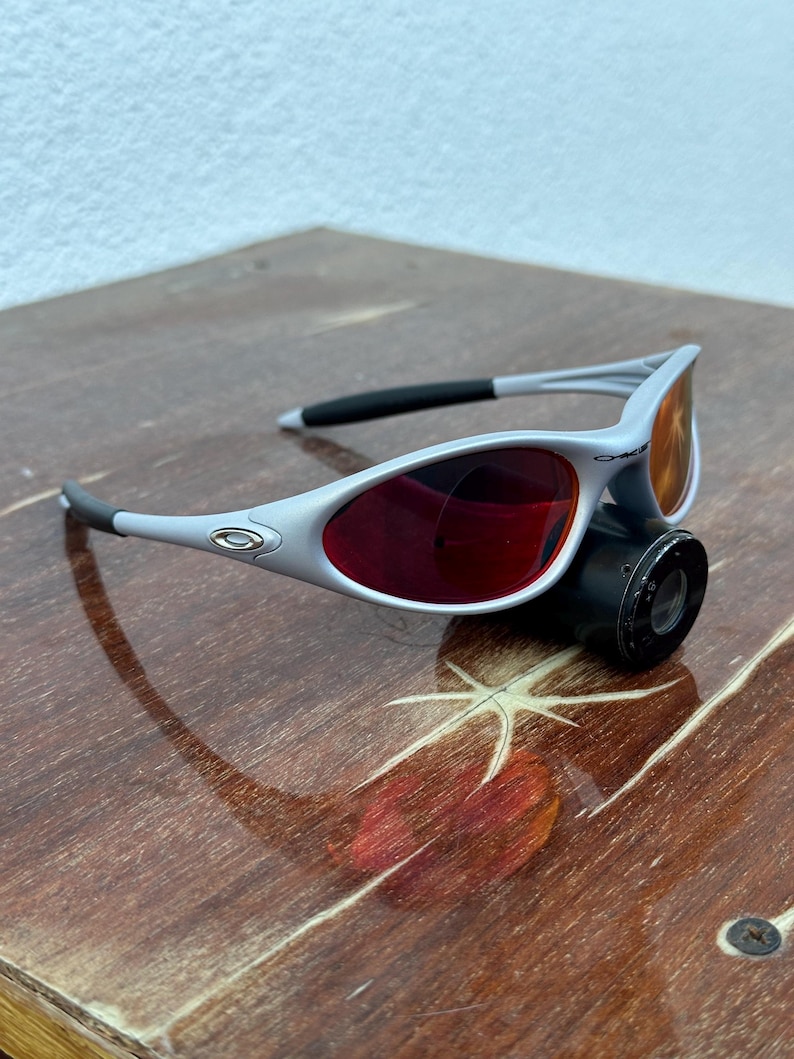 May include: Gray and black sunglasses with red lenses. The sunglasses have a sleek, modern design with a silver logo on the temples. The lenses are a vibrant red color, and the frames are a light gray.