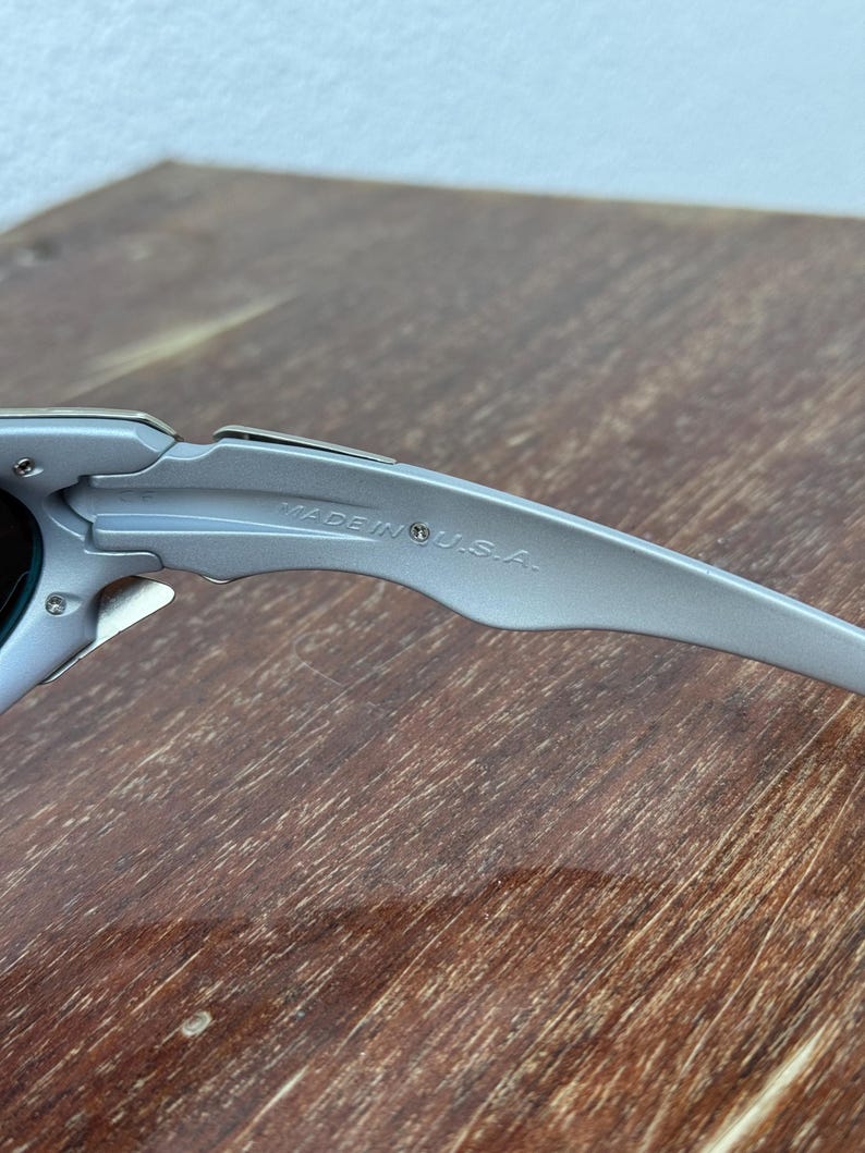 Puede incluir: Primer plano de unas gafas de sol plateadas con una patilla curvada. El texto "MADE IN U.S.A." est&aacute; grabado en la patilla. Las gafas de sol est&aacute;n sobre una superficie de madera.