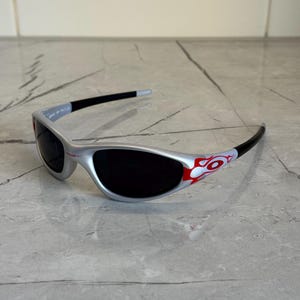 May include: Silver sunglasses with black lenses and black arms. The arms feature a red and white flame design. The sunglasses are on a gray surface.