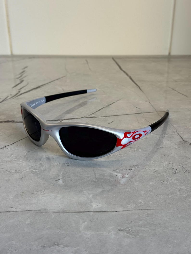 May include: Silver sunglasses with black lenses and black arms. The arms feature a red and white flame design. The sunglasses are on a gray surface.