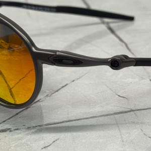 Pu&ograve; includere: Primo piano di un paio di occhiali da sole con montatura grigio scuro e lenti arancione-giallo. Gli occhiali hanno un design elegante con una montatura sottile e aste curve. Le lenti hanno un effetto sfumato.