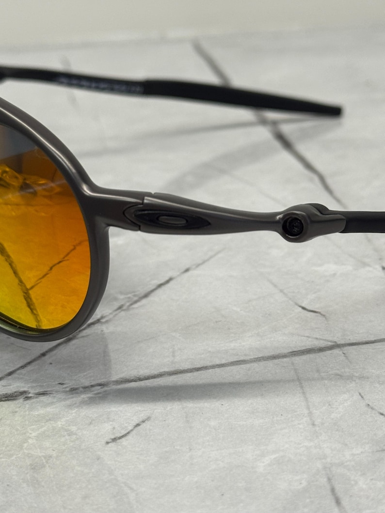 Pu&ograve; includere: Primo piano di un paio di occhiali da sole con montatura grigio scuro e lenti arancione-giallo. Gli occhiali hanno un design elegante con una montatura sottile e aste curve. Le lenti hanno un effetto sfumato.