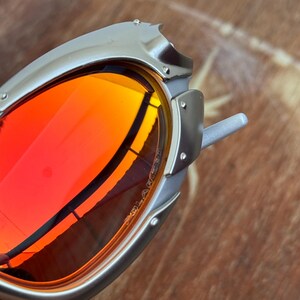 Puede incluir: Primer plano de gafas de sol con montura plateada y lentes rojas vibrantes. Las gafas de sol tienen un dise&ntilde;o angular &uacute;nico con remaches visibles y una est&eacute;tica elegante y moderna. Las lentes reflejan el entorno.