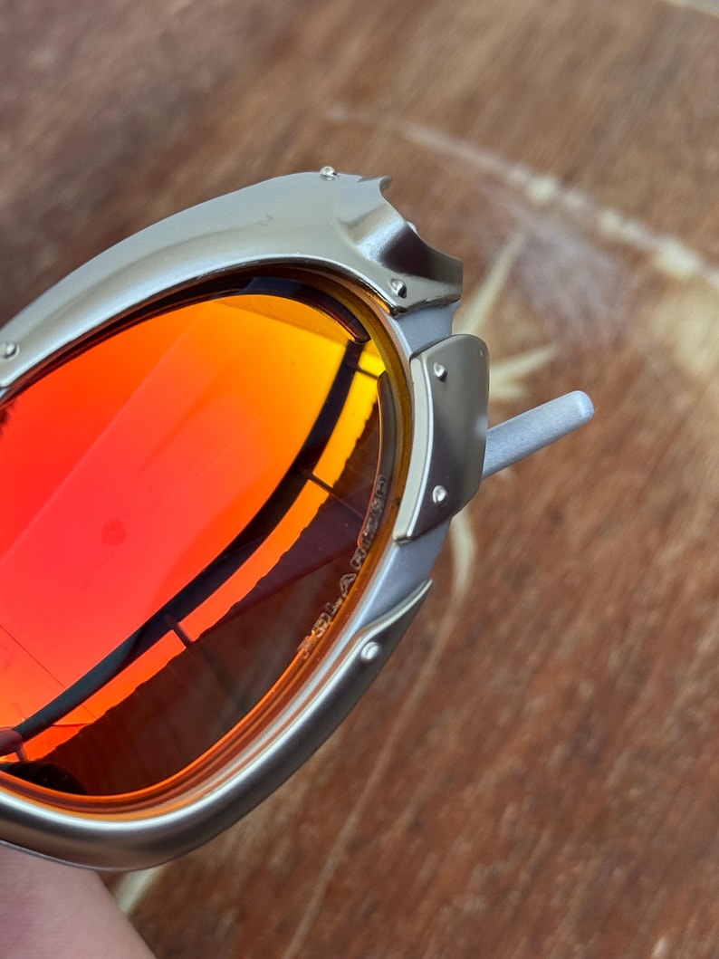 Puede incluir: Primer plano de gafas de sol con montura plateada y lentes rojas vibrantes. Las gafas de sol tienen un dise&ntilde;o angular &uacute;nico con remaches visibles y una est&eacute;tica elegante y moderna. Las lentes reflejan el entorno.