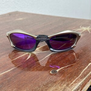 May include: A pair of silver-toned sunglasses with purple lenses. The sunglasses have a curved, wraparound design.