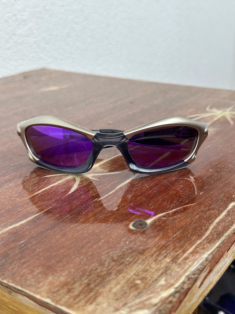 May include: A pair of silver-toned sunglasses with purple lenses. The sunglasses have a curved, wraparound design.