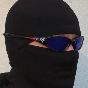 May include: Black and red framed sunglasses with blue lenses. The sunglasses have a star design on the side and are worn with a black balaclava. The balaclava covers the head and neck.