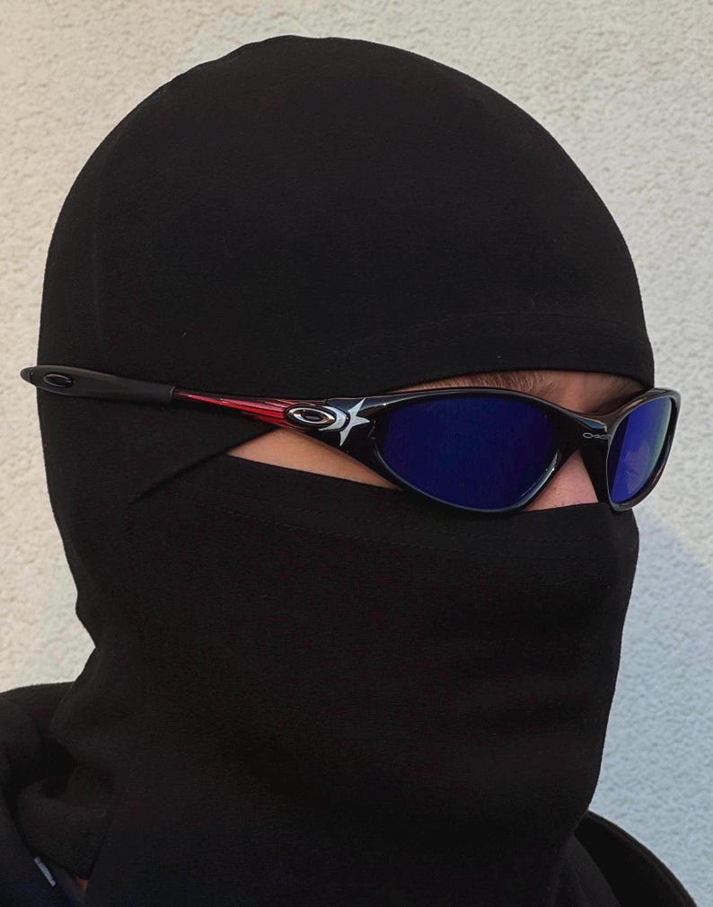 May include: Black and red framed sunglasses with blue lenses. The sunglasses have a star design on the side and are worn with a black balaclava. The balaclava covers the head and neck.