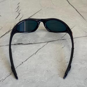 May include: Black sunglasses with dark green lenses. The sunglasses have a curved frame and arms, designed for a secure fit. The lenses are a dark green color, offering protection from the sun. The sunglasses are likely designed for sports or outdoor activities.