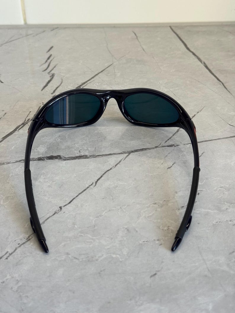 May include: Black sunglasses with dark green lenses. The sunglasses have a curved frame and arms, designed for a secure fit. The lenses are a dark green color, offering protection from the sun. The sunglasses are likely designed for sports or outdoor activities.