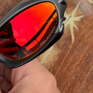 Op de afbeelding: Zwarte zonnebril met rode gepolariseerde glazen. De glazen reflecteren de omgeving. Het montuur is matzwart. Het woord "POLARIZED" is zichtbaar op het glas.