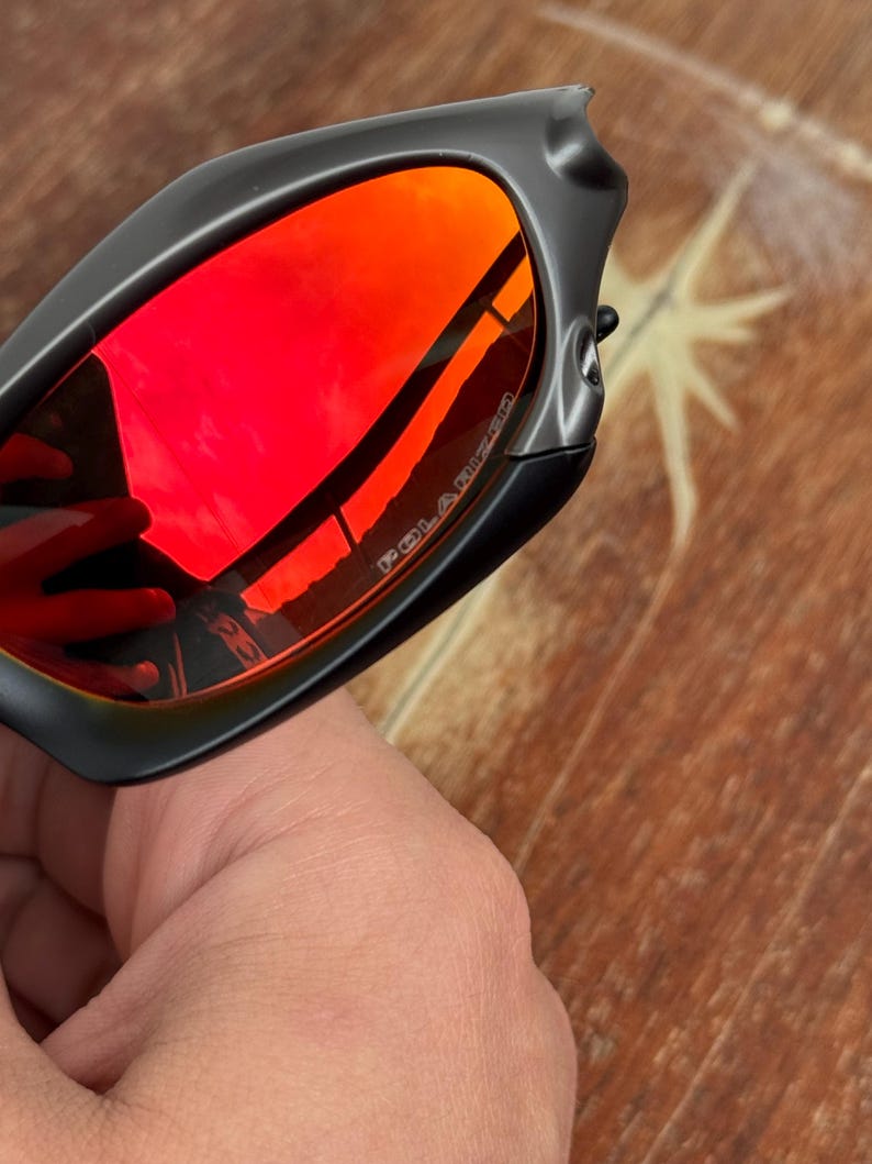 Op de afbeelding: Zwarte zonnebril met rode gepolariseerde glazen. De glazen reflecteren de omgeving. Het montuur is matzwart. Het woord "POLARIZED" is zichtbaar op het glas.