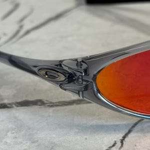 K&ouml;nnte beinhalten: Nahaufnahme einer Sonnenbrille mit transparentem grauem Rahmen und leuchtend roten Gl&auml;sern. Das Oakley-Logo ist auf dem B&uuml;gel zu sehen. Die Sonnenbrille befindet sich vor einem hellen, strukturierten Hintergrund.